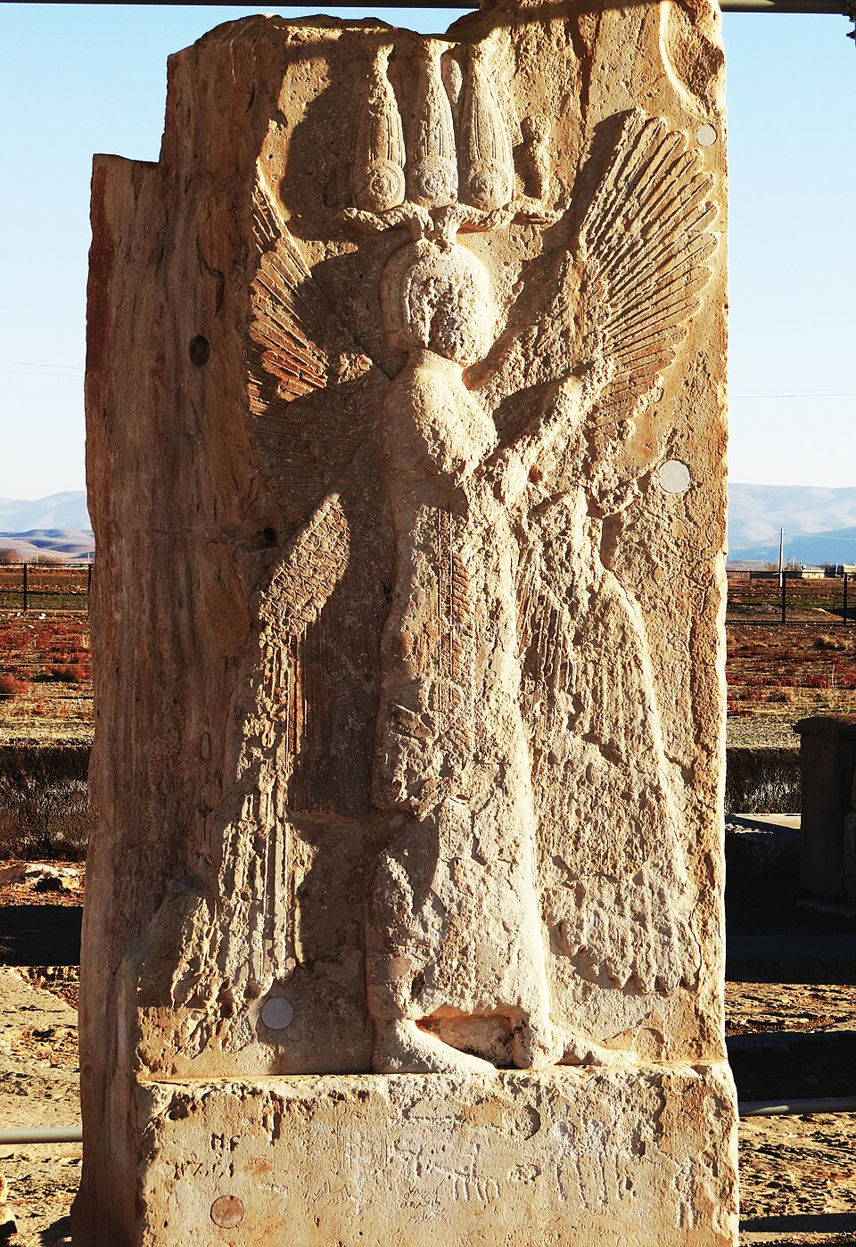 Relief de l'homme ailé à Parsagades, Iran.
Description longue : une grande stèle de pierre ocre, assez abîmée, montrant un homme marchant de profil. Quatre ailes partent du corps de l'homme. Il porte une tunique d'aspect assez rigide et une sorte de lourd couvre-chef avec une structure portant trois coiffes longues, comme s'il portait trois fois la couronne des pharaons égyptiens (?).