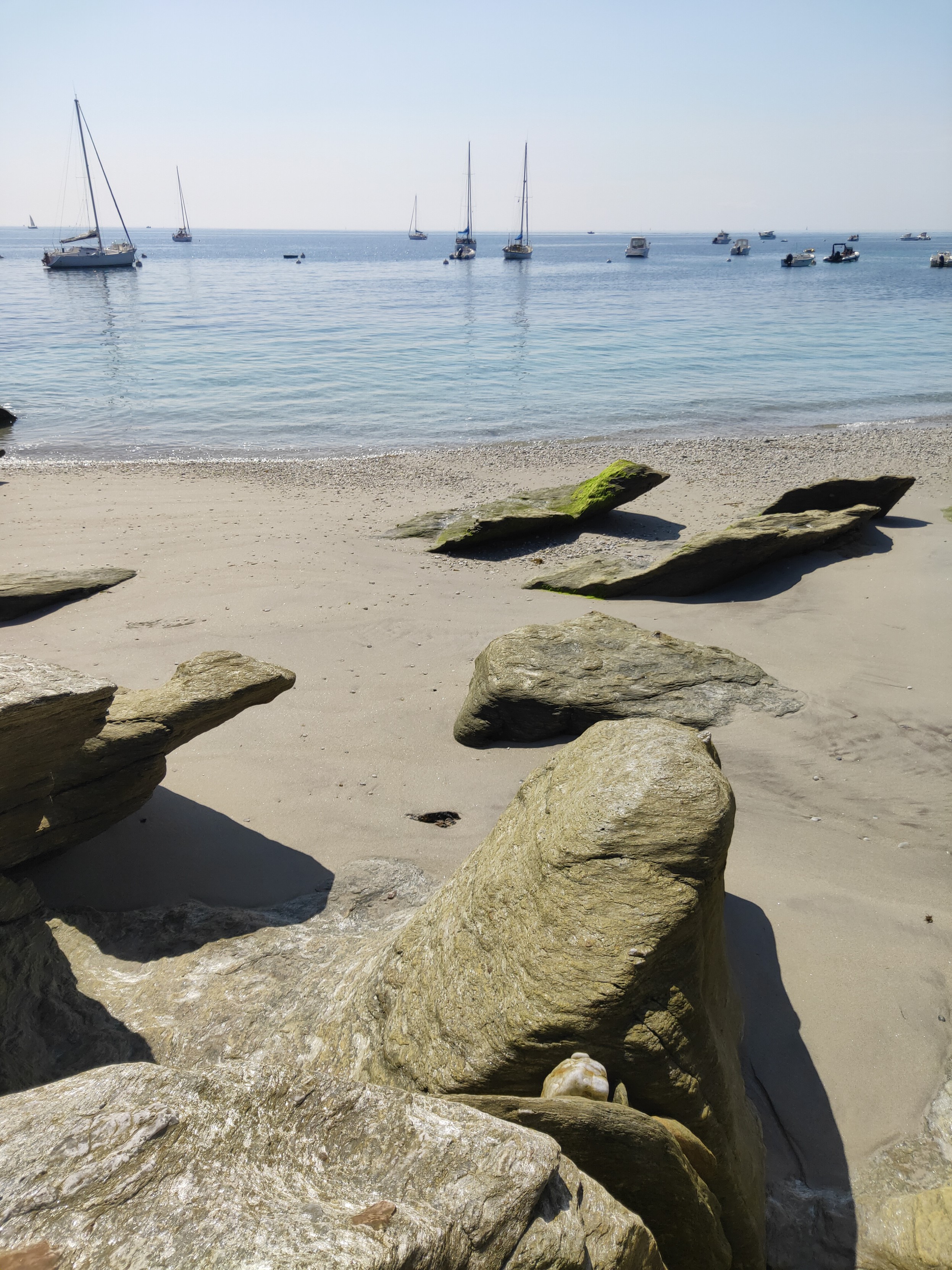 Au premier plan, quelques rochers exposés plein soleil, plus ou moins proéminents et aux différentes formes qui émergent du sable, certains sont recouverts à certains endroits d'une fine mousse verte. Au second plan, une mer calme d'une couleur bleu turquoise et un peu plus loin, plusieurs bateaux amarrés dont des voiliers sous un ciel bleu pâle.