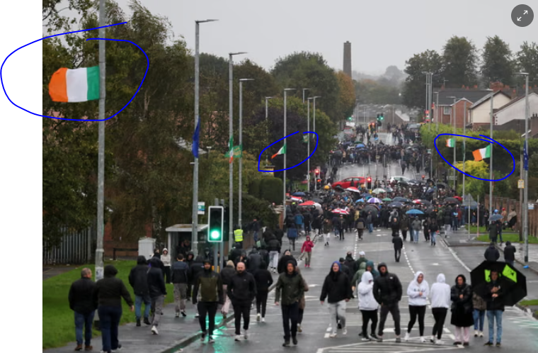 Photo de Garvaghy Road, rue de Portadown, sur le Guardian. On voit une rue par temps de pluie, avec des groupes de gens habillés de noir et tenant des parapluies. Des drapeaux irlandais flottent à des mâts.